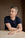 A man with short gray hair in a navy t-shirt sits at a wooden table, looking thoughtfully to the side.