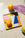 A geometric-patterned plate with an orange sits on fabric, surrounded by yellow and green fruits and a purple mesh bag.