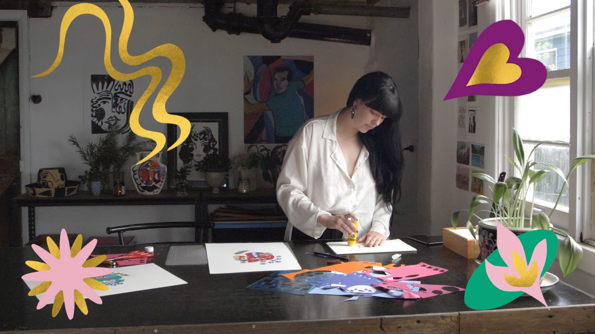 A woman in a white shirt crafts at a table, surrounded by colorful paper art in a bright, cozy studio.