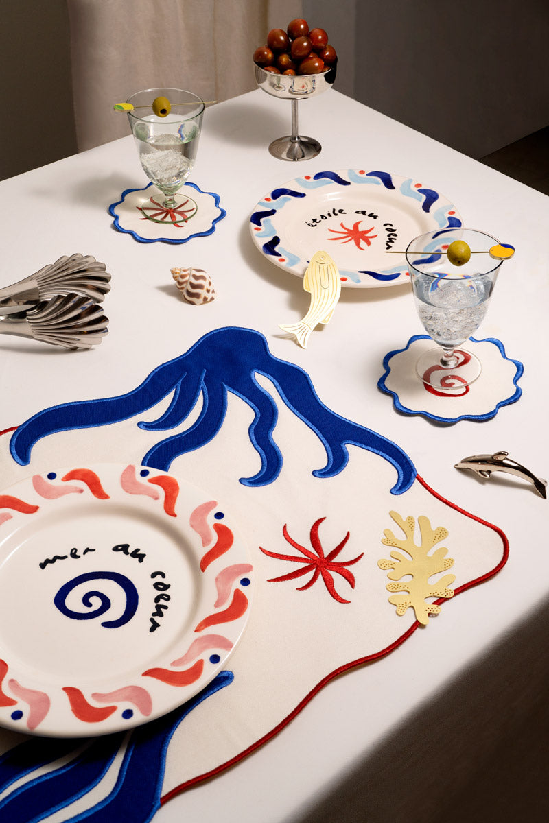 A table set with artistic ocean-themed plates, wavy placemats, shell décor, and glasses garnished with olives.