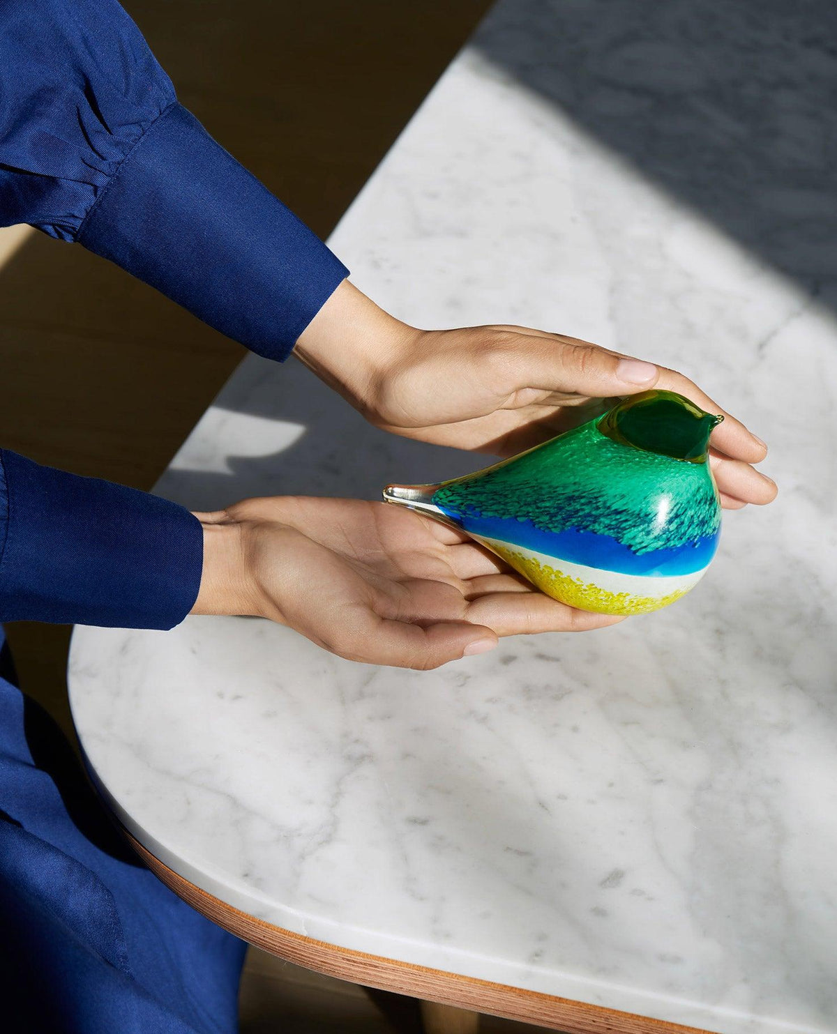 Someone in a blue sleeve holds the blue OCTAEVO Atlas Glass Bird, a vibrant decor piece, on a marble table.