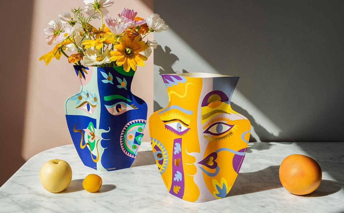 Two colorful vases with abstract face designs, one holding yellow and white flowers, sit on a marble surface with fruits