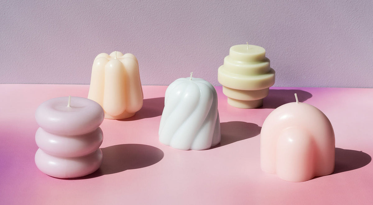 Quatre bougies décoratives aux teintes pastel, aux formes géométriques originales, sont disposées devant un fond rose clair.