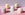 Four pastel-colored decorative candles in unique geometric shapes are arranged against a light pink background.