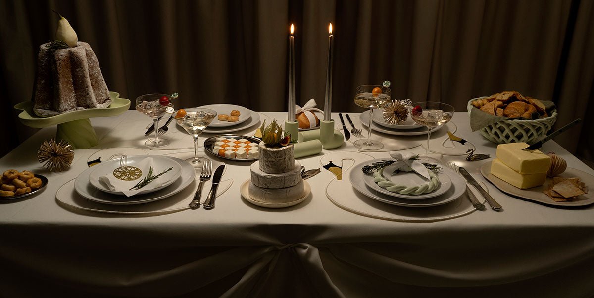 A dinner table set for four with dishes, two lit candles, drinks, a small cake, pastries, bread, butter, and holiday accents.