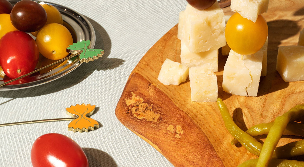 A wooden board with cheese and green peppers are next to a plate of tomatoes. Pasta-shaped cocktail picks are placed nearby.