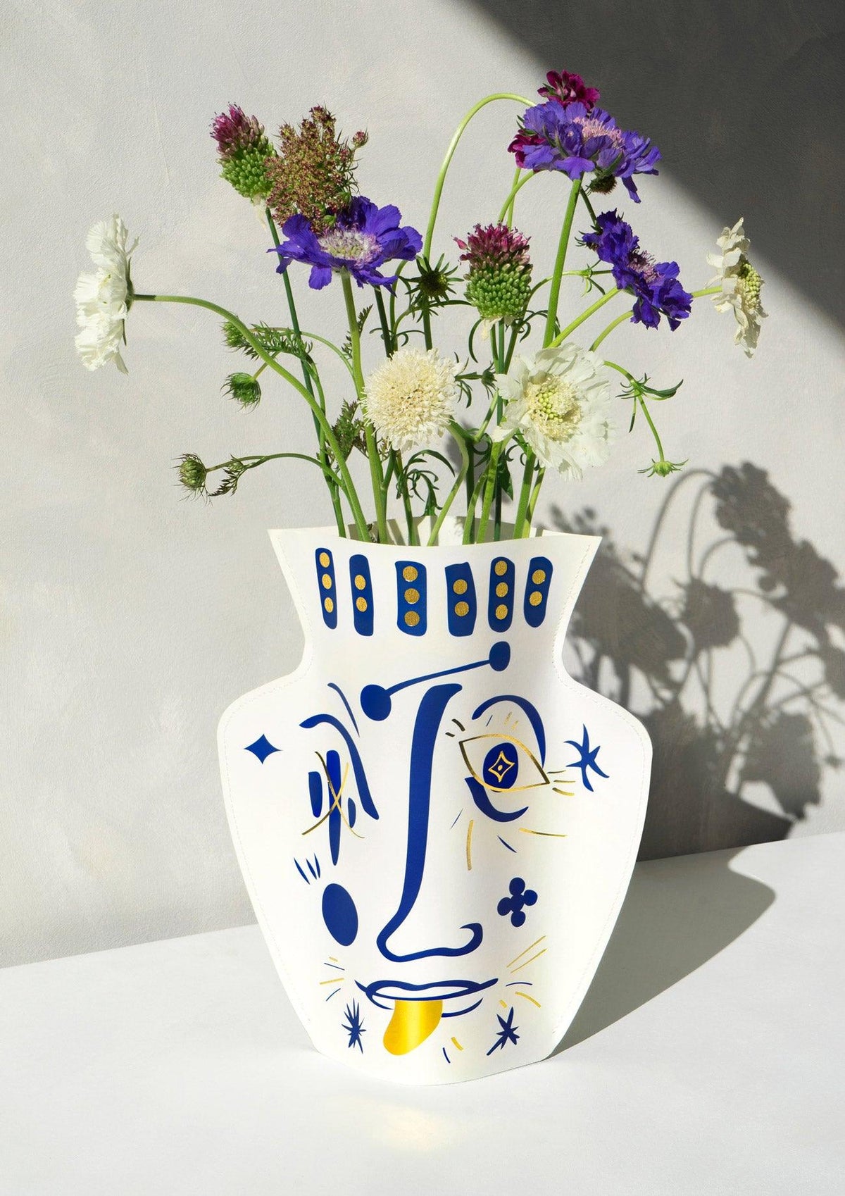 OCTAEVO Jaime Hayon Paper Vase — Vasage White with abstract blue-yellow face, sunlit and filled with wildflowers.