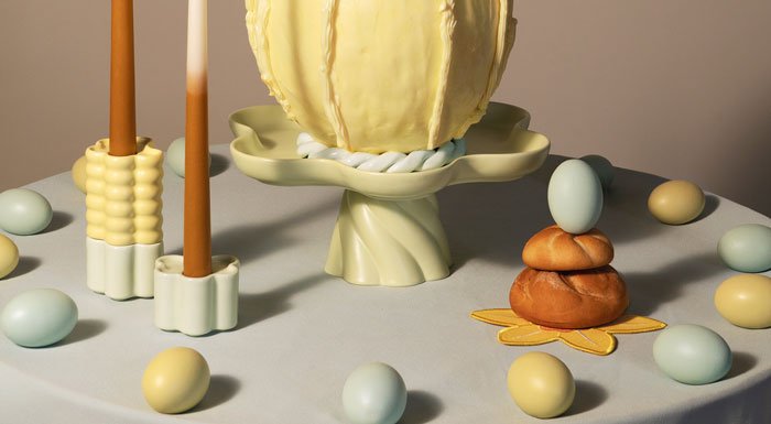 A table displays pastel eggs, a yellow butter egg on a stand, tall candles in ceramic holders, and a stack of bread rolls.
