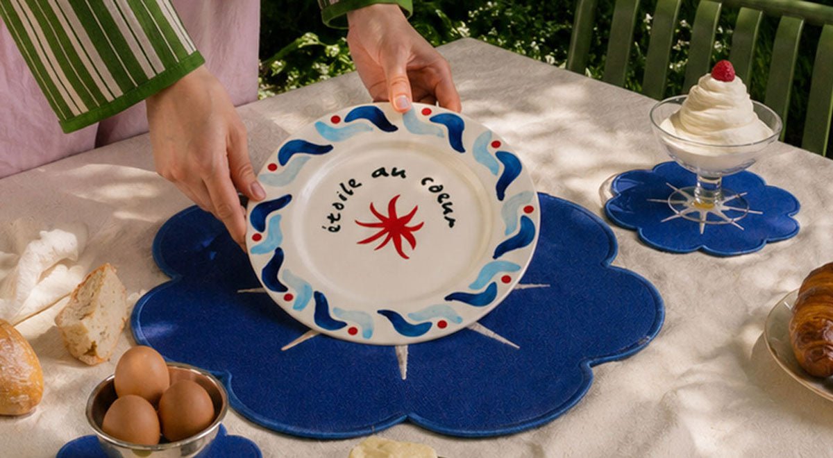 A person holds a decorative plate with text, above a blue flower placemat on a table set with breakfast.