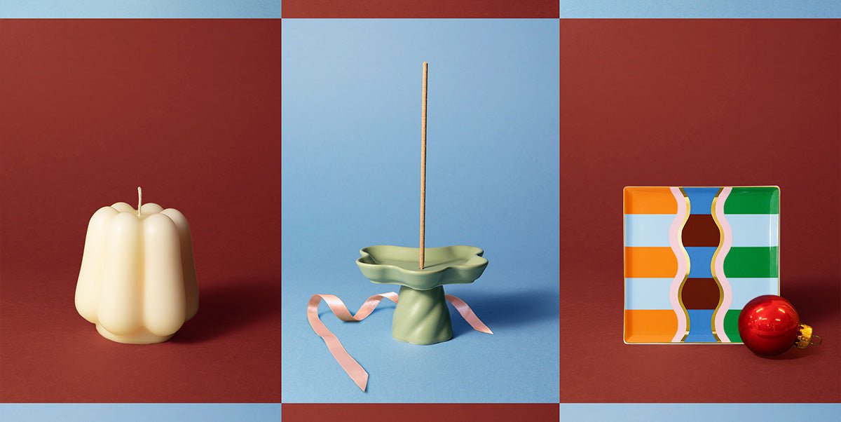 A white candle, a green wavy incense holder with a stick and pink ribbon, and striped tray, on red and blue backgrounds.