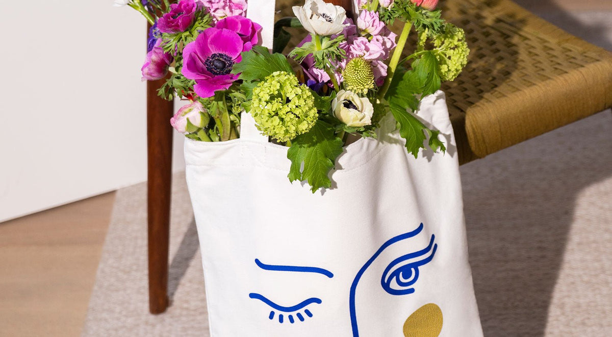A white tote bag with a minimalist blue face design holds a bouquet of colorful flowers, resting on a chair.