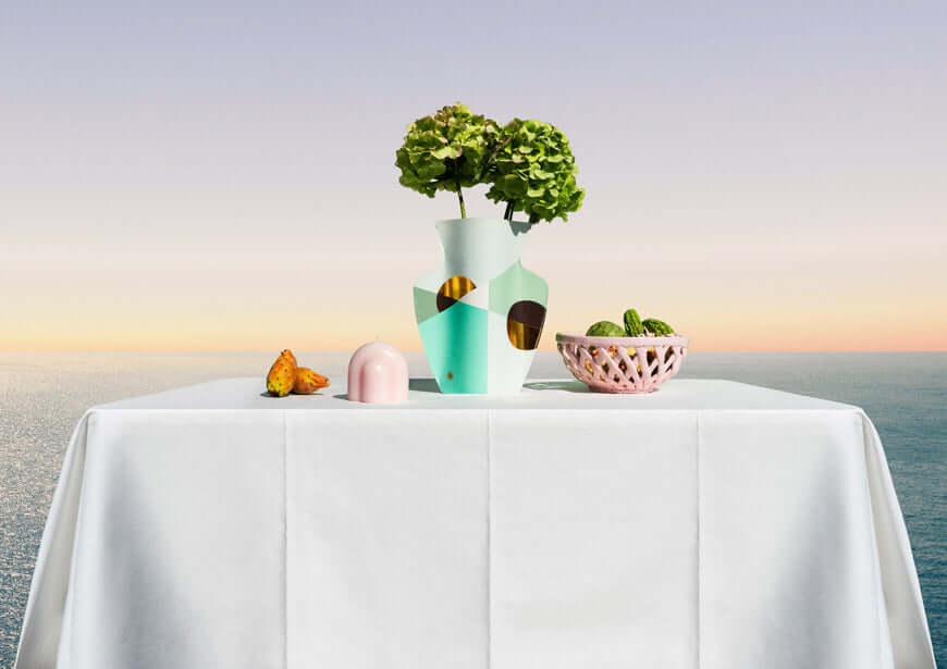 A table with a white cloth is set outdoors by the sea at sunset, featuring a modern vase with hydrangeas, a pink dome object, a woven bowl of cucumbers, and a cut fig.