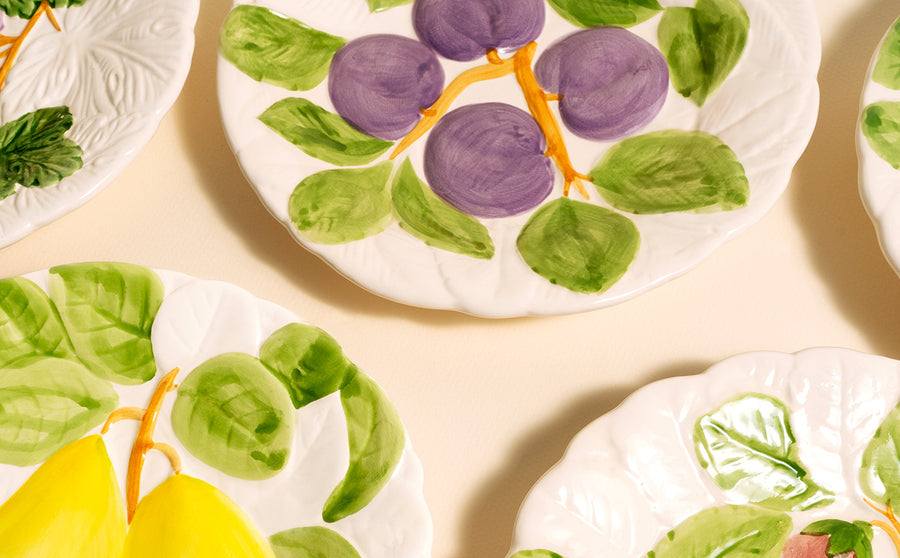 Colorful ceramic plates with hand-painted fruit designs, including purple plums and yellow pears, on a cream background.