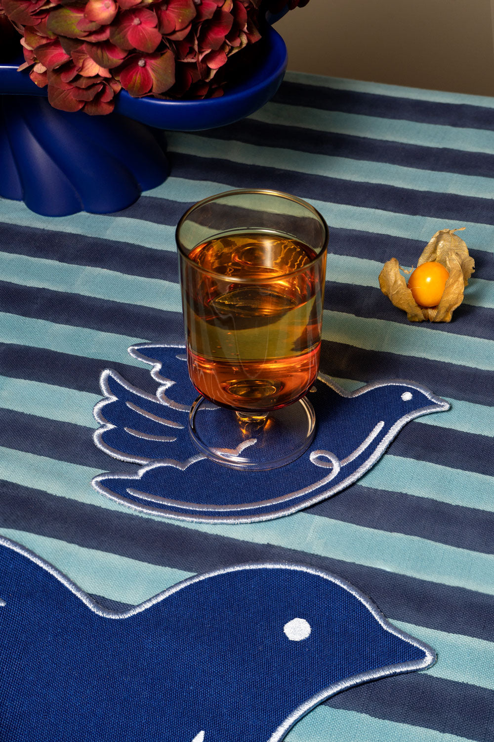 A clear glass tumbler filled with amber liquid sits on a blue bird-shaped placemat, placed on a striped tablecloth.