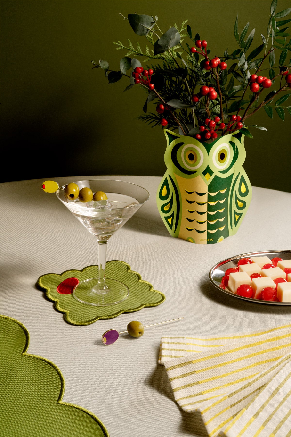 Mini vase en papier Night Owl par OCTAEVO avec des baies, un verre à martini, un plateau de fromages et une serviette rayée, sur une table verte et blanche.