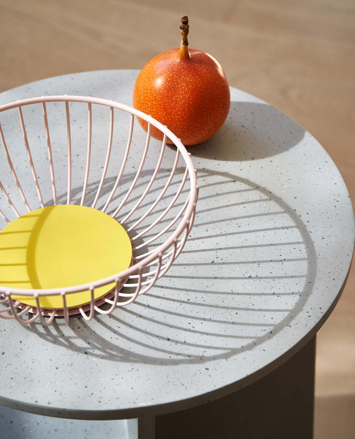 A pink wire fruit basket with a yellow base sits on a round table next to an orange passion fruit.