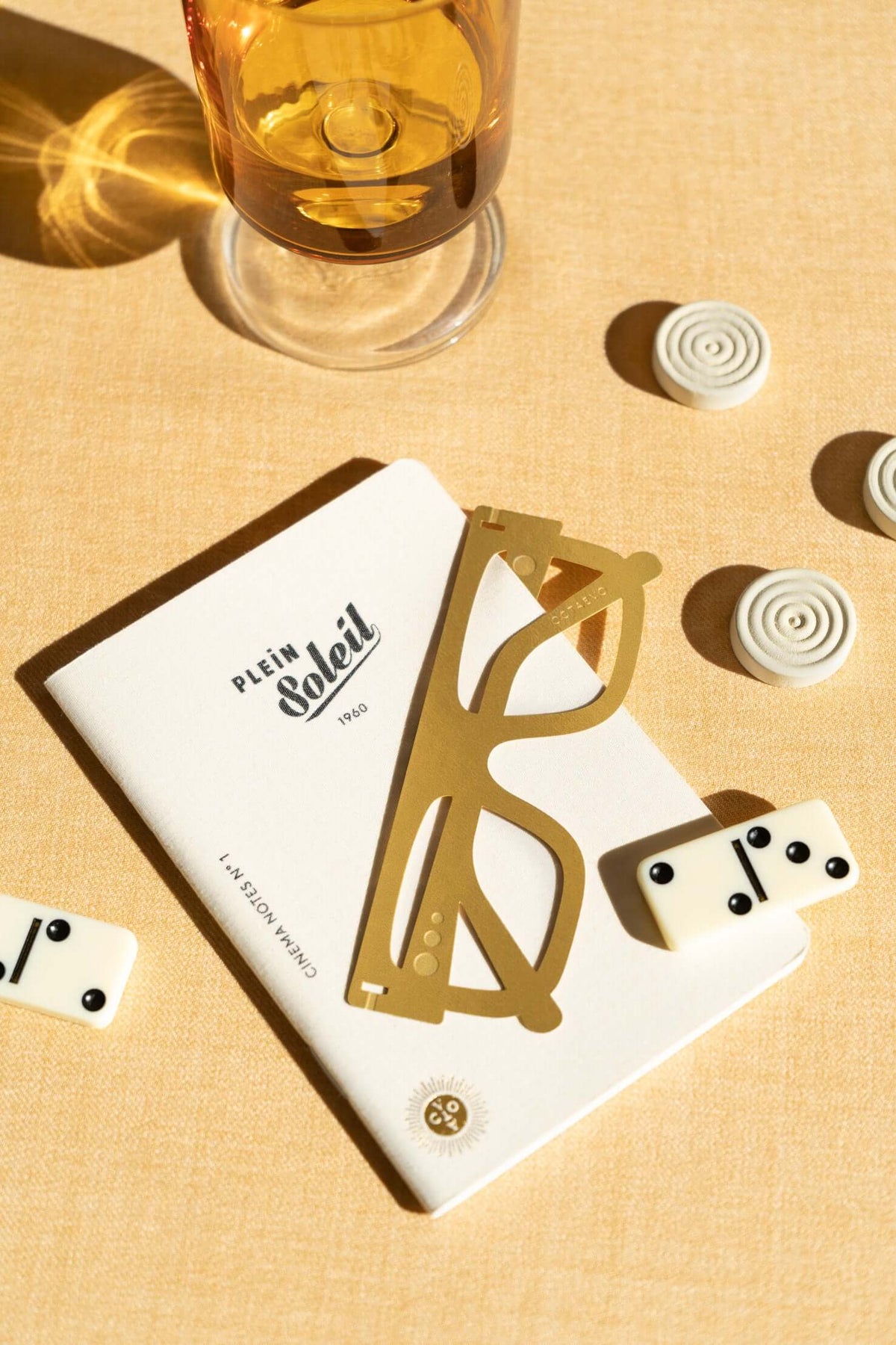 A glass of drink, dominoes, a notebook, and gold glasses placed on a beige surface in bright sunlight.