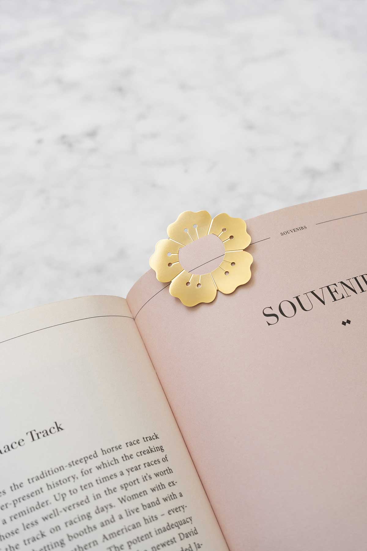 A gold flower-shaped bookmark sits on the edge of an open book on a marble surface.
