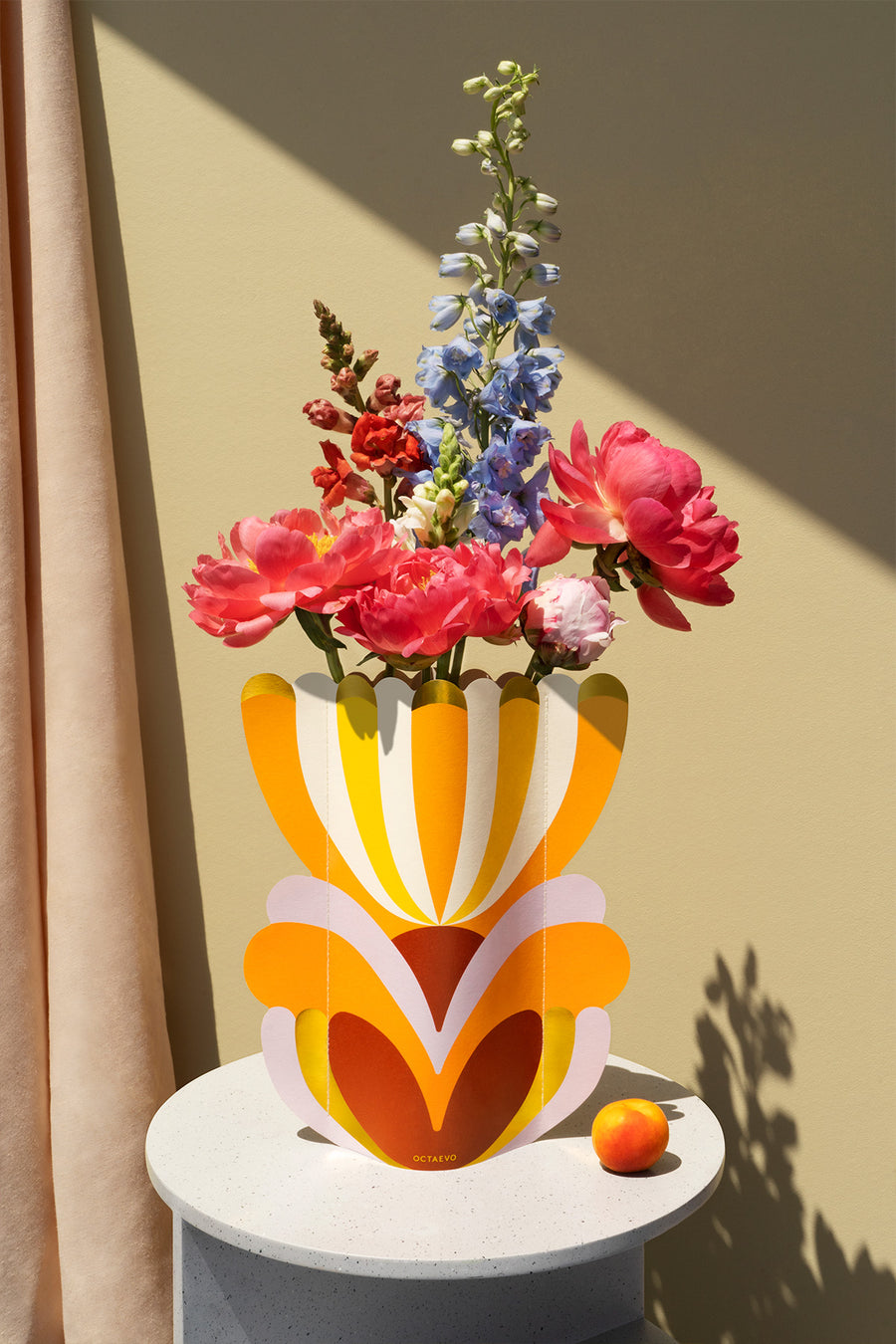 A colorful vase with bright flowers and a small peach on a round table against a pale yellow wall and peach curtain.
