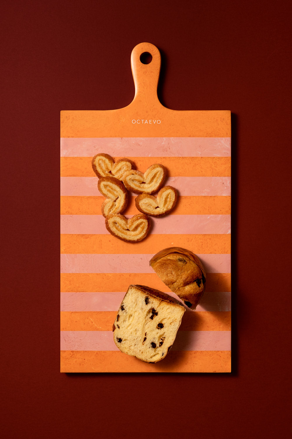 An orange and pink striped wooden cutting board with a handle, styled with heart-shaped cookies and a sliced loaf of bread, set against a solid dark red background. The brand name 'OCTAEVO' is printed on the board.