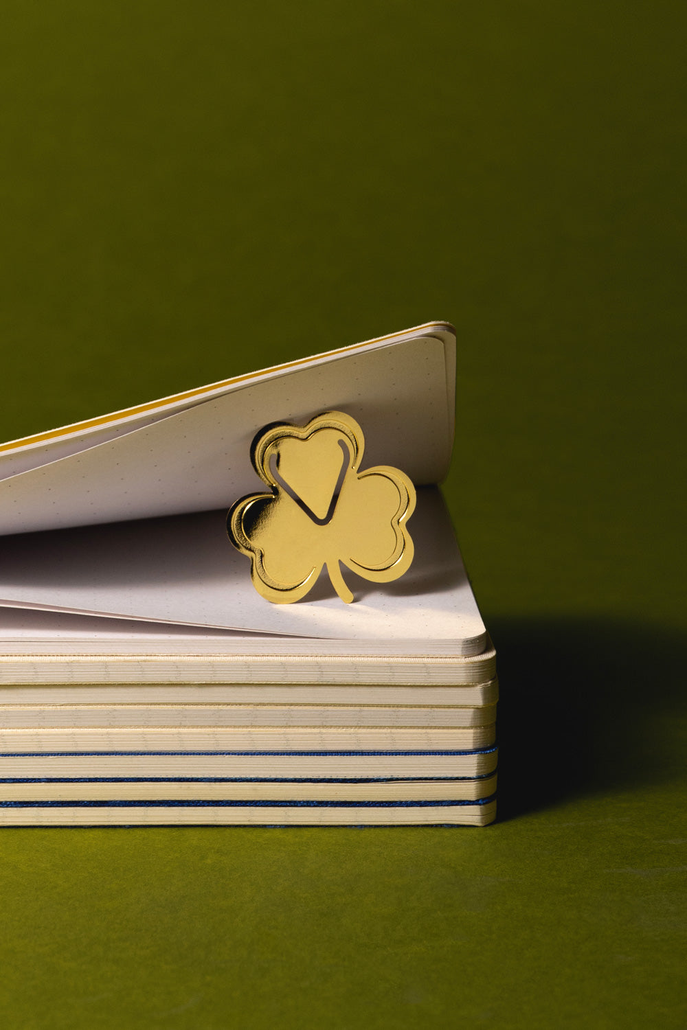A gold metallic shamrock bookmark resting on the open pages of a stack of white notebooks against a solid olive green background.