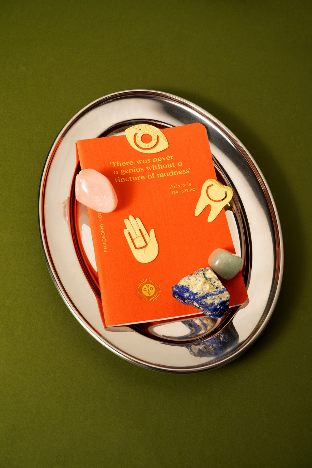 An orange book or booklet with gold illustrations and text lies on a silver oval tray, surrounded by small polished stones against an olive green background.