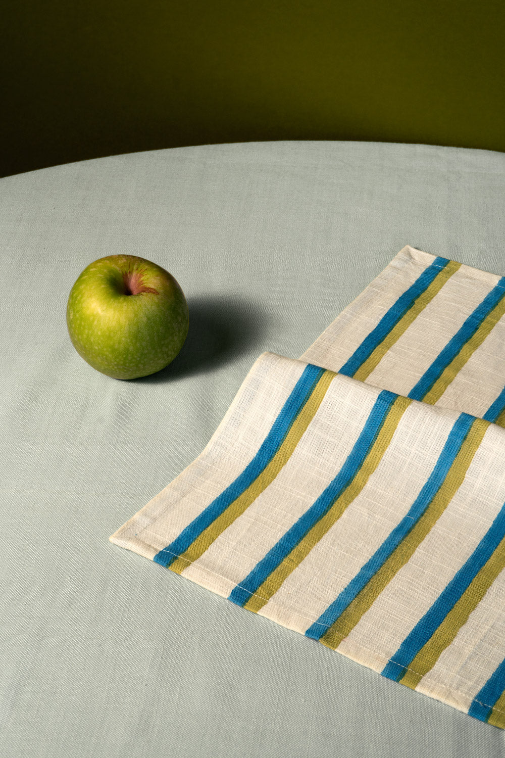 Eine gefaltete Leinenserviette mit Längsstreifen in Blau und Olivgrün, arrangiert auf einer hellgrauen Tischdecke neben einem Apfel.
