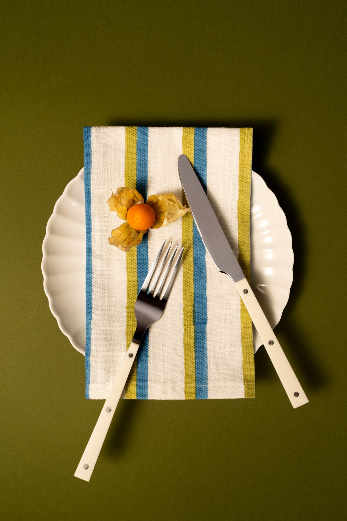 Flatlay-Ansicht eines Gedecks mit einem weißen Keramikteller, einer gestreiften Leinenserviette mit senkrechten Streifen in Blau und Olivgrün, einem silbernen Messer mit cremefarbenem Griff und einer passenden silbernen Gabel mit cremefarbenem Griff, alles auf einem einfarbig olivgrünen Hintergrund arrangiert.