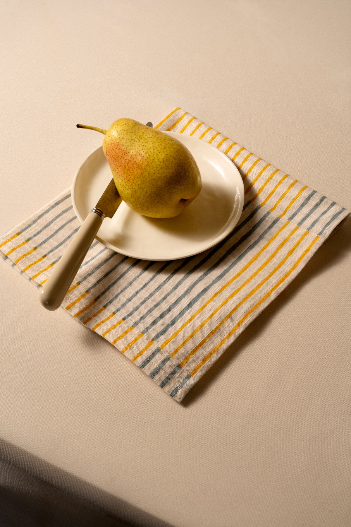 Una pera reposa sobre un pequeño plato blanco con un cuchillo de mango de madera, colocado sobre una servilleta de tela a rayas naranjas, blancas y azules, en una mesa beige.