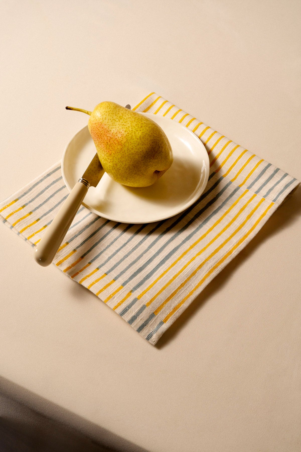 Riviera napkin in Orange, under a small plate with a pear and a wooden-handled knife.