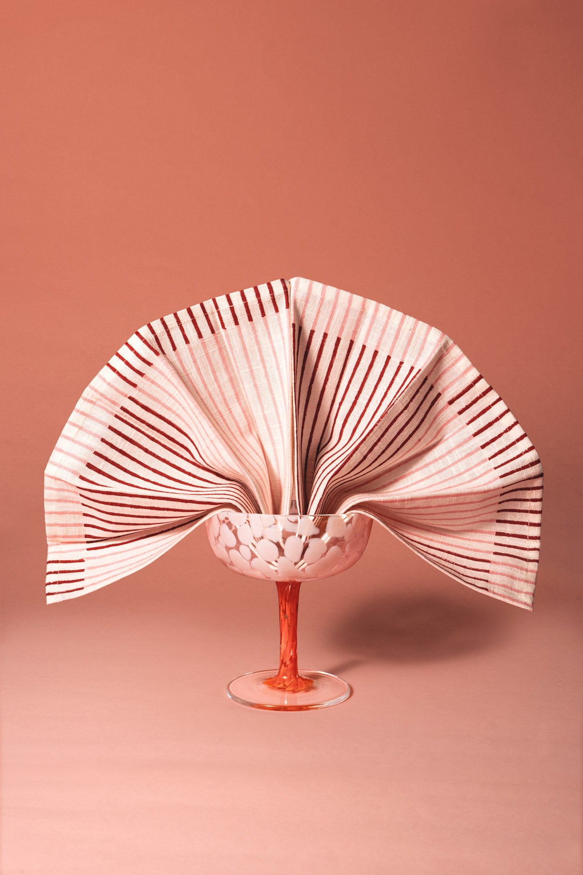 Una escultura de papel decorativa en forma de abanico, con rayas rosas y blancas, exhibida sobre un pedestal de vidrio transparente contra un fondo rosa empolvado.