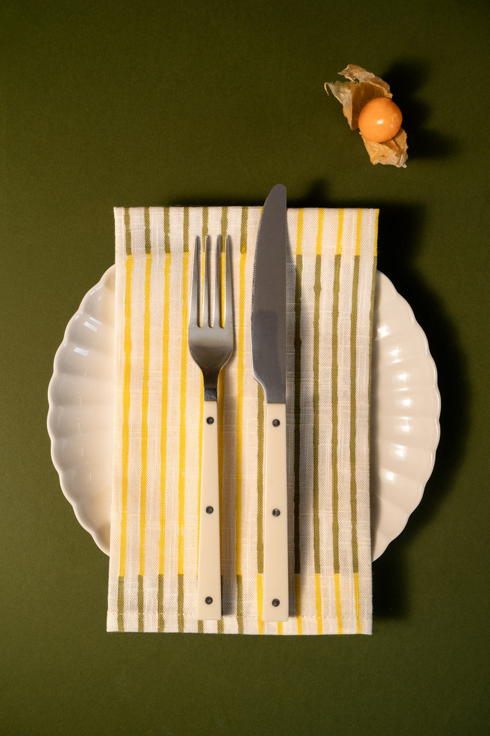 Una vista cenital de una mesa con una servilleta a rayas verticales amarillas y verde oliva, un plato blanco festoneado, un cuchillo plateado y un tenedor plateado con mangos de madera clara. Una pequeña fruta en su cáscara de papel se encuentra en la esquina superior derecha, sobre un fondo verde oliva oscuro.