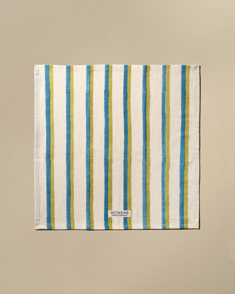 A square cotton napkin with vertical stripes in blue, featuring a small white brand label at the bottom that reads 'OCTAEVO'. The towel is displayed flat against a solid beige background.