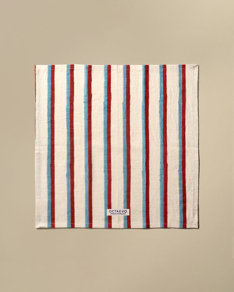 A square cotton napkin with vertical stripes in red, featuring a small white brand label at the bottom that reads 'OCTAEVO'. The towel is displayed flat against a solid beige background.