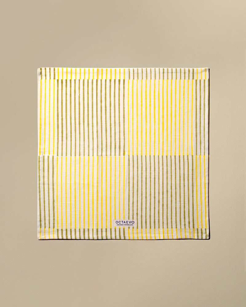 A square cotton napkin with stripes in yellow, featuring a small white brand label at the bottom that reads 'OCTAEVO'. The towel is displayed flat against a solid beige background.