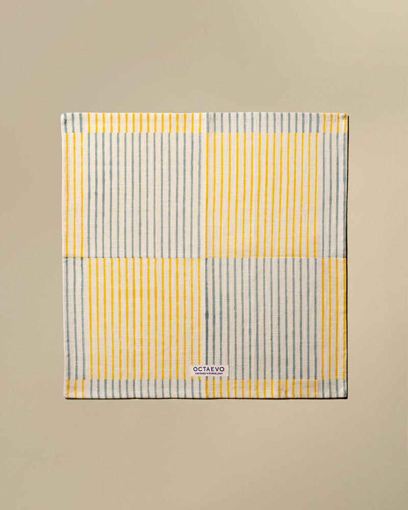 A square cotton napkin with stripes in orange, featuring a small white brand label at the bottom that reads 'OCTAEVO'. The towel is displayed flat against a solid beige background.