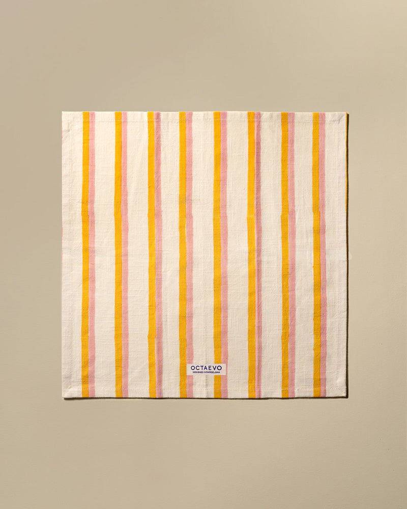 A square cotton napkin with vertical stripes in orange, featuring a small white brand label at the bottom that reads 'OCTAEVO'. The towel is displayed flat against a solid beige background.