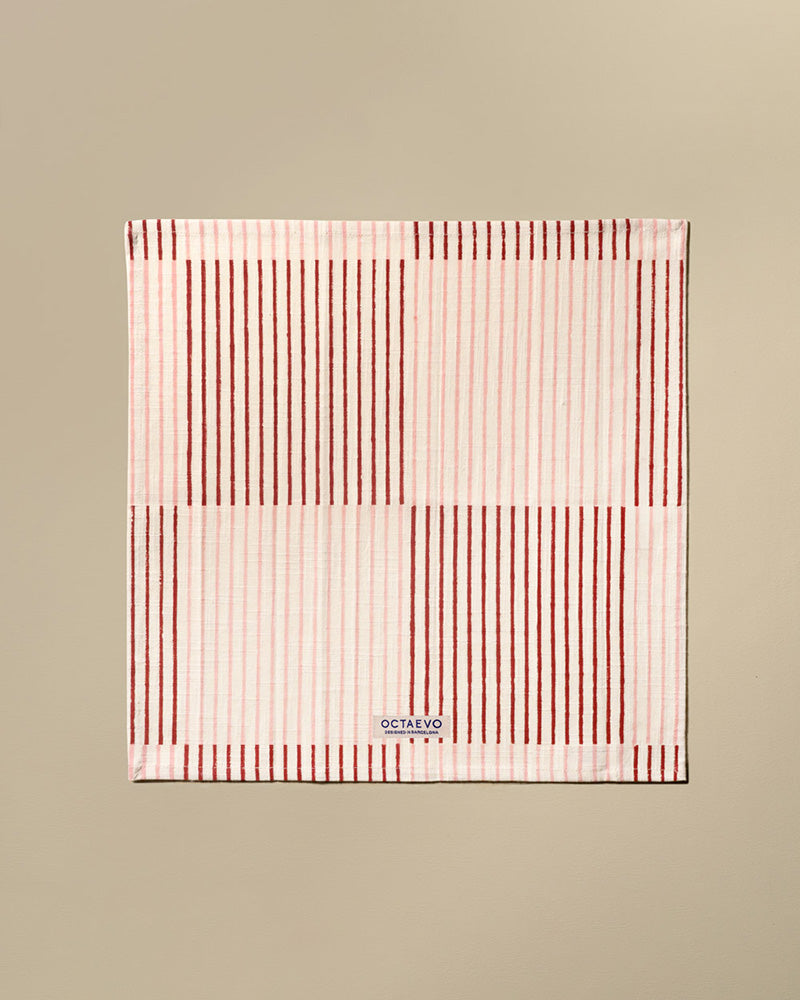 A square cotton napkin with stripes in pink, featuring a small white brand label at the bottom that reads 'OCTAEVO'. The towel is displayed flat against a solid beige background.