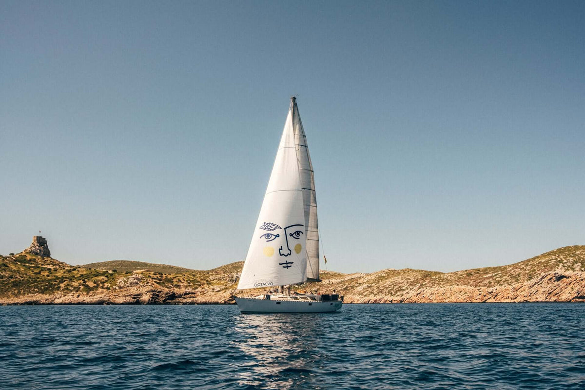 A sailboat with a face drawn on its sail glides on blue water near rocky, green hills under a clear sky.