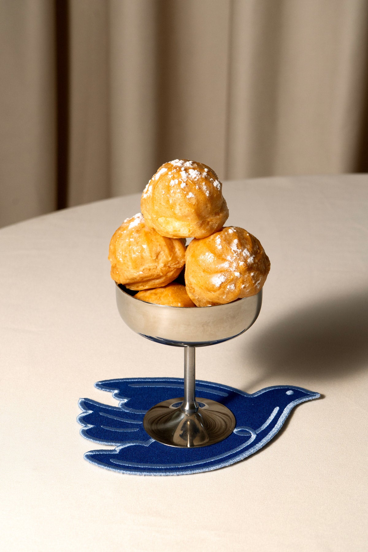 Three golden-brown choux pastries on a silver pedestal stand, placed on a blue bird-shaped trivet on a light table with a curtain background.