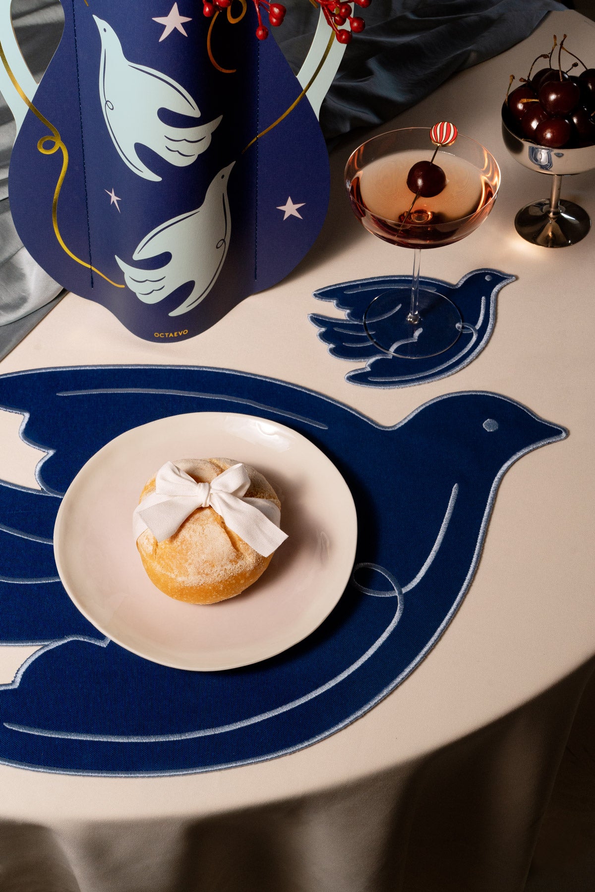 A styled tabletop scene featuring a large   bird-shaped placemat in blue with white linework, a small white plate holding a pastry tied with a white bow, a blue decorative pitcher, a cocktail glass, and a bowl of cherries.