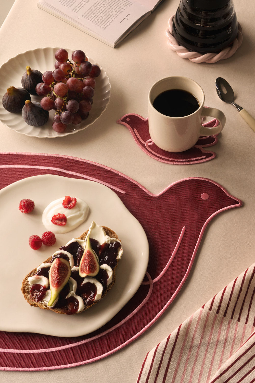 A red bird-shaped placemat styled on a warm beige table, with a cream plate holding fig and berry toast, a coffee cup on a matching bird-shaped coaster, fresh grapes and figs, and a striped napkin.