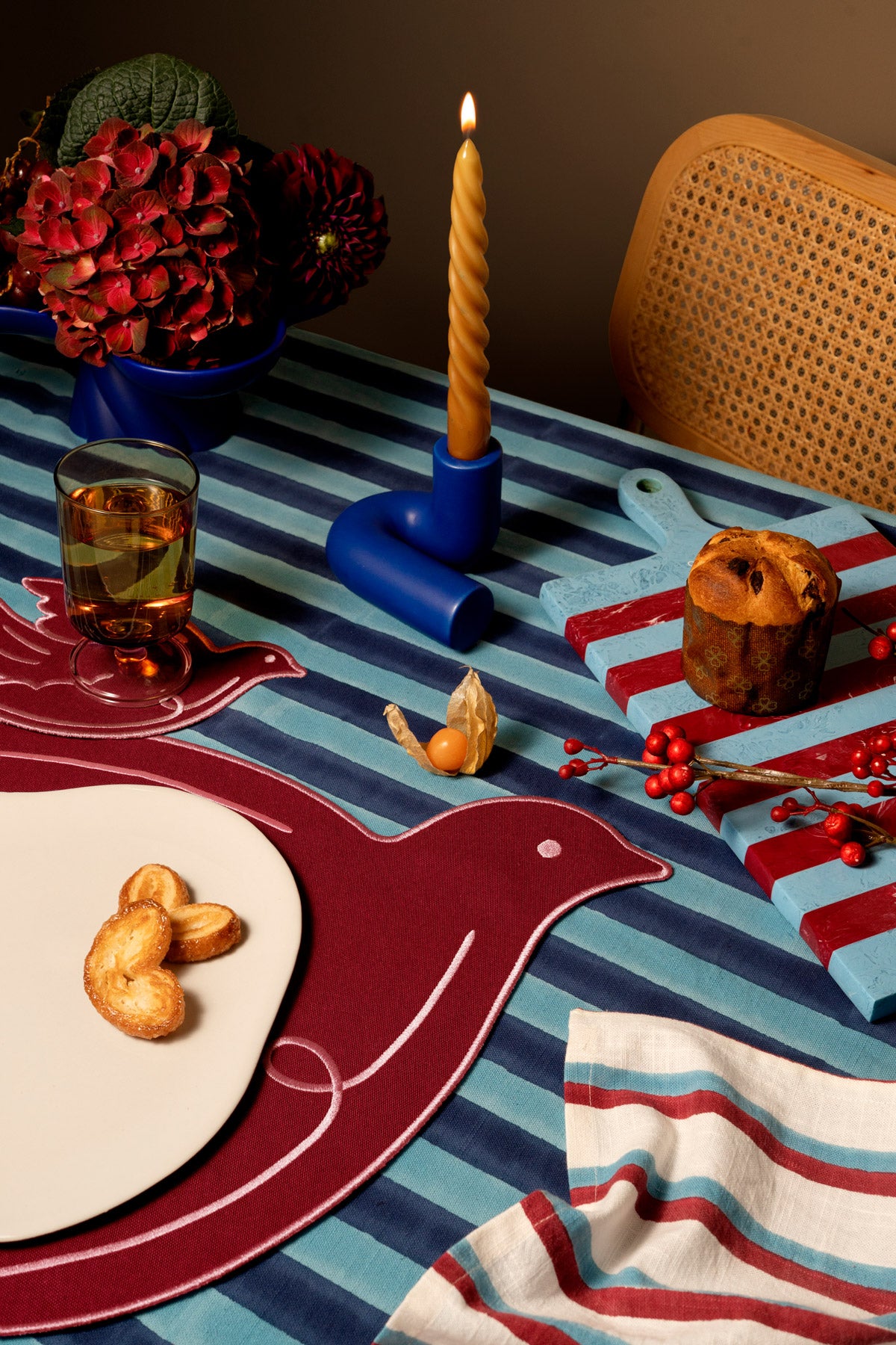 A styled dining table setting striped tablecloth,  bird-shaped placemat in red, a striped napkin, a candle holder, a glass of amber drink, and decorative food items.