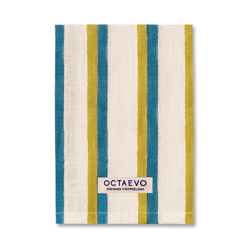 Eine rechteckige Serviette mit senkrechten Streifen in Blau, Senfgelb und Creme, mit einem Markenetikett am unteren Rand mit der Aufschrift 'OCTAEVO DESIGNED IN BARCELONA'.