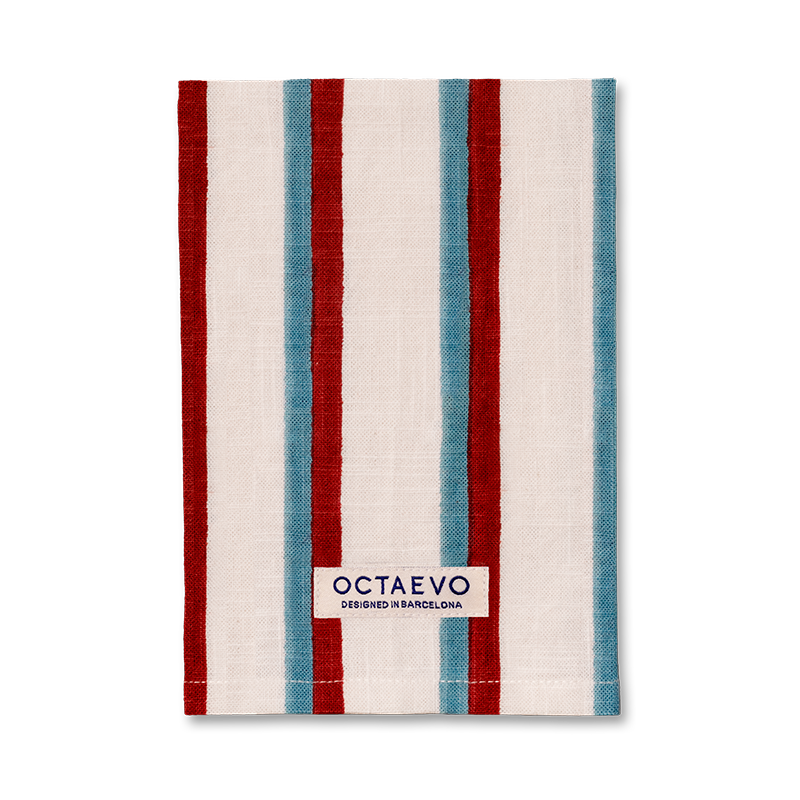 Eine rechteckige Serviette mit Längsstreifen in Blau, Rot und Creme, unten mit einem Markenetikett mit der Aufschrift 'OCTAEVO DESIGNED IN BARCELONA'.
