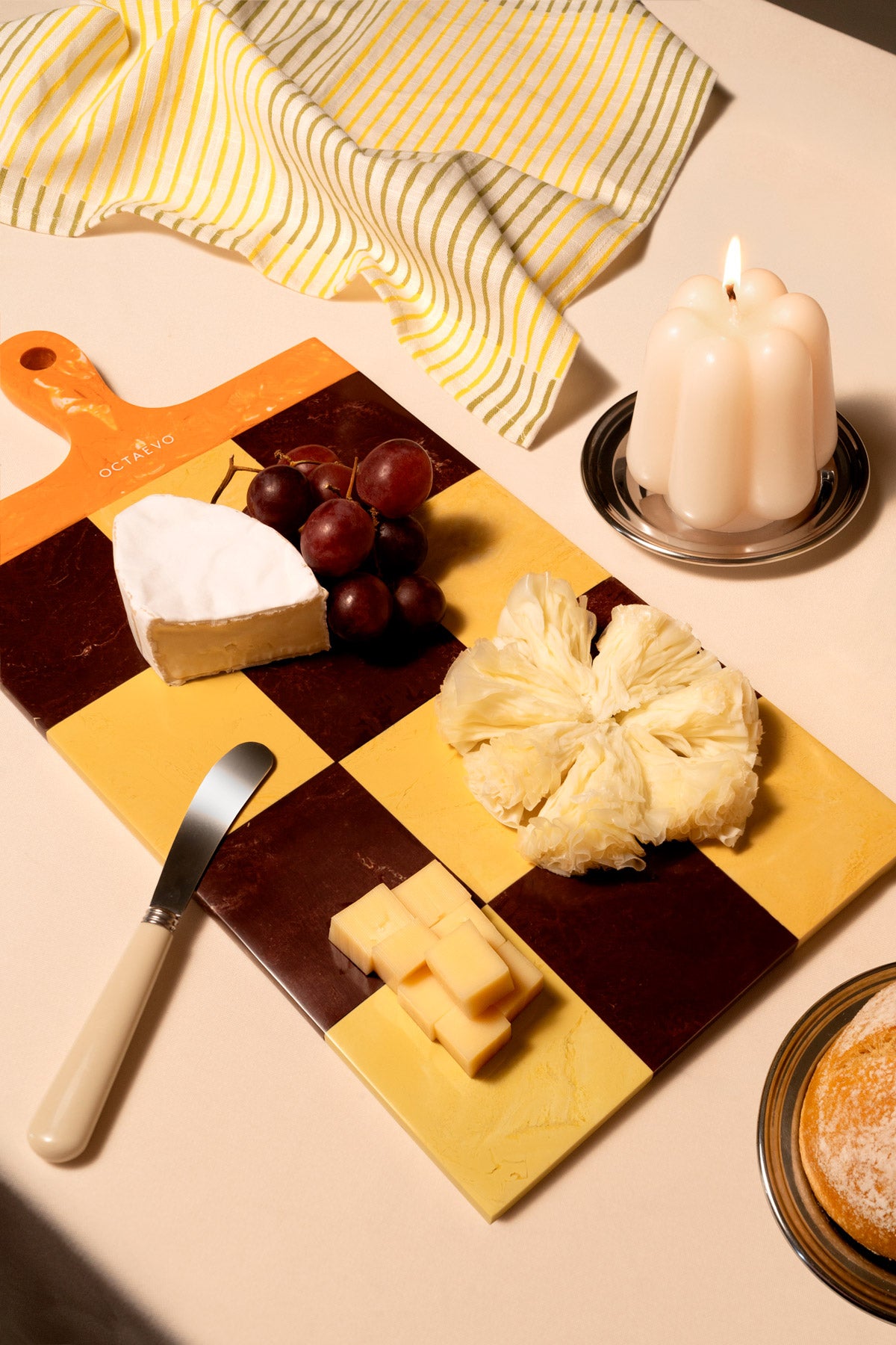 A checkered wooden cheese board with yellow and dark brown squares, styled with various cheeses, grapes, a knife, a striped cloth, and a lit candle on a beige table.
