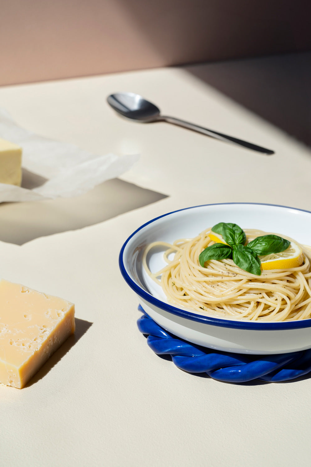 A white ceramic bowl with a blue rim filled with spaghetti, garnished with fresh green basil leaves and a slice of lemon, set on a blue textured trivet. The scene is styled with blocks of cheese and a silver spoon on a beige surface under warm, direct sunlight.