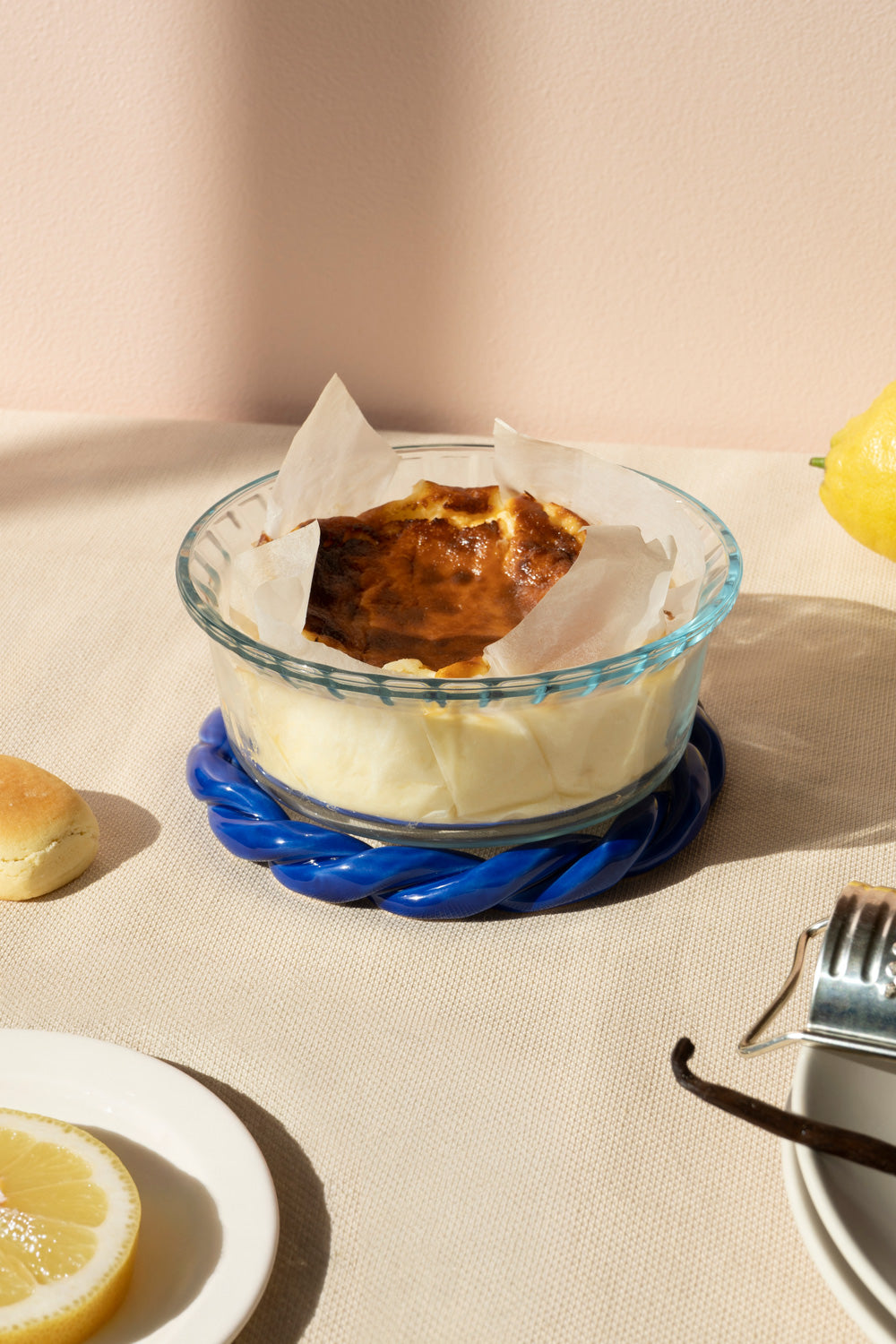 A clear glass baking dish with a blue twisted trivet, containing a pale cream-colored dessert or cake topped with a browned custard-like layer and parchment paper, styled on a beige tablecloth with lemon slices and a small pastry nearby.