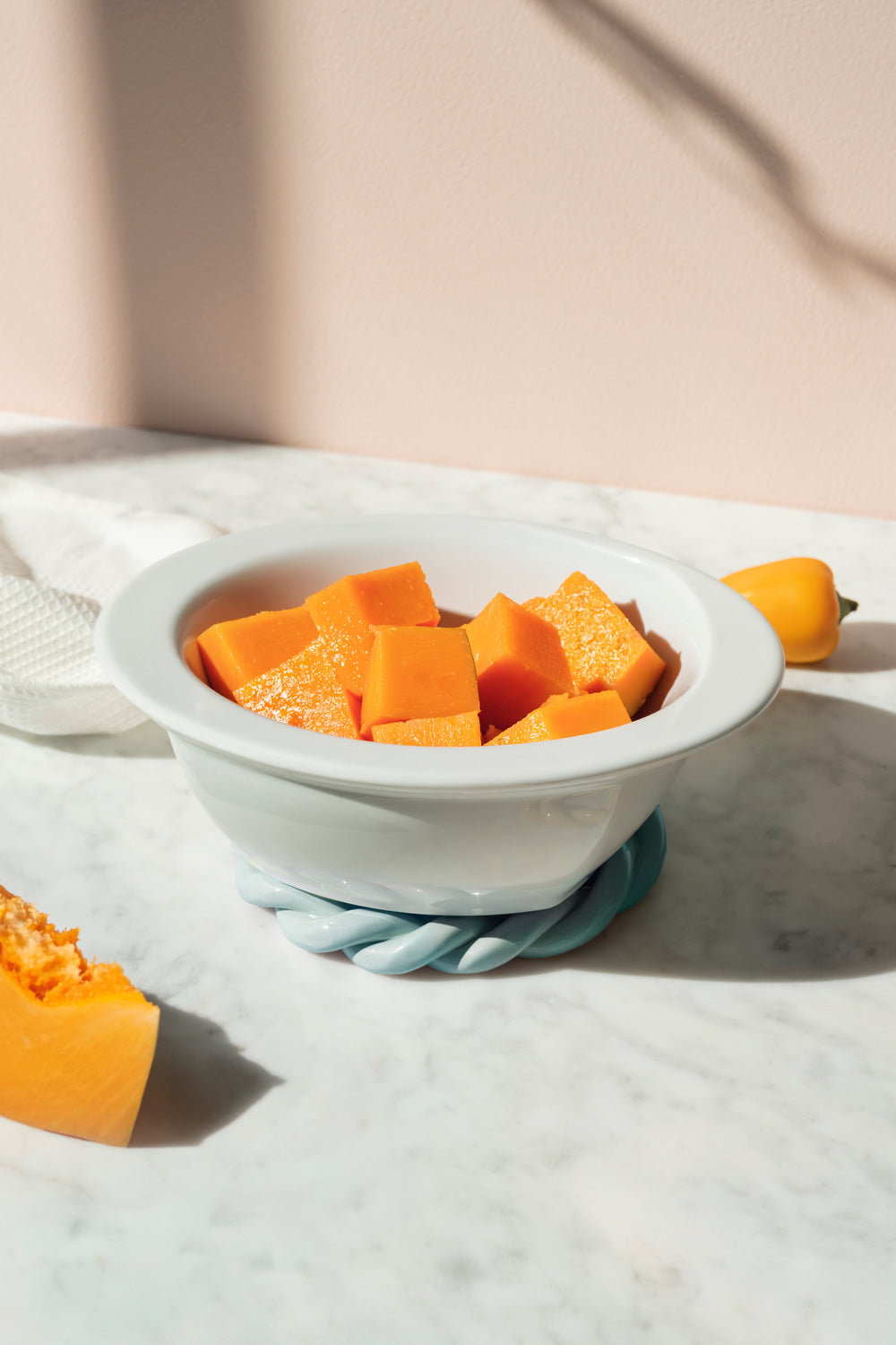 A white ceramic bowl filled with cubed pumpkin or squash, set on a light marble surface with a light blue trivet. Sunlight casts shadows on a soft beige wall in the background.