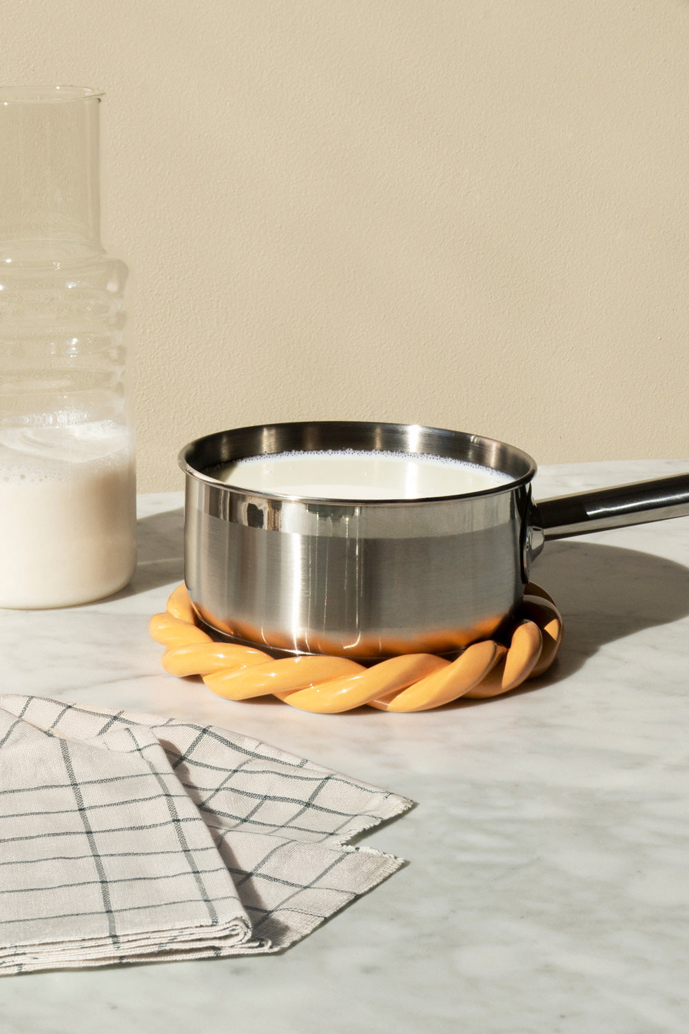 A stainless steel saucepan filled with white milk sits on a twisted orange trivet on a light marble surface, with a glass bottle of milk and a checkered cloth in the background.